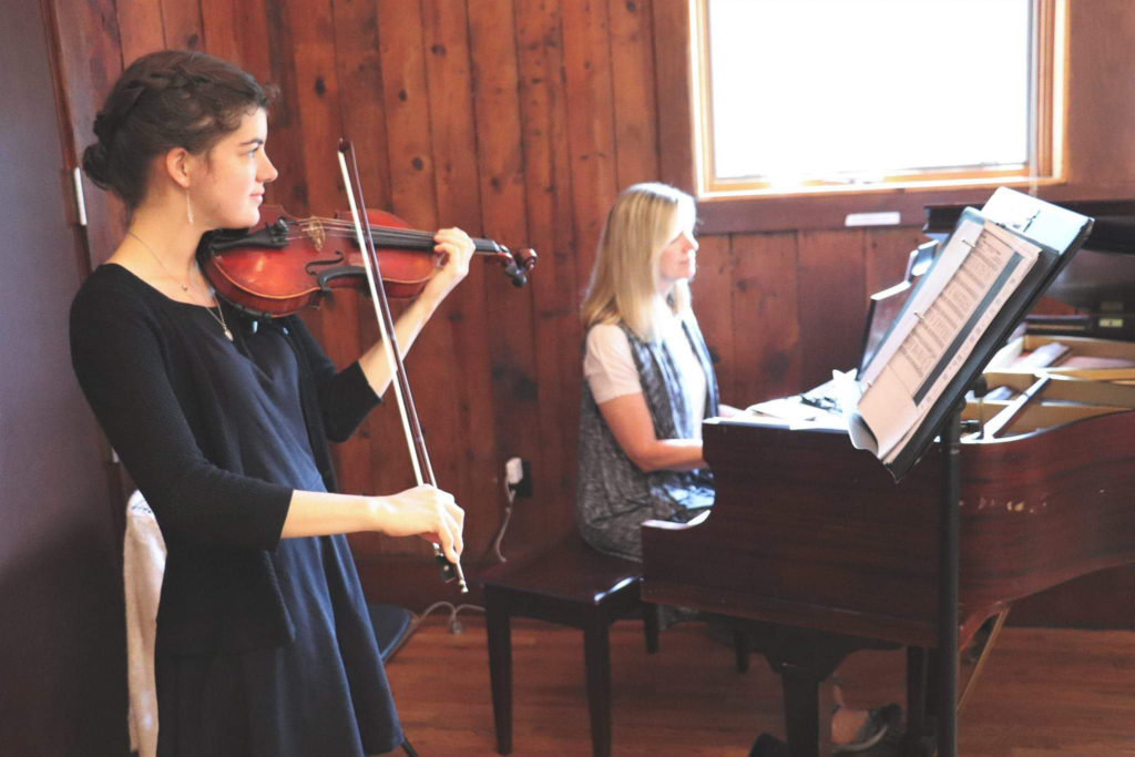 A girl plays violin at a wedding event, while a women plays piano in the background.